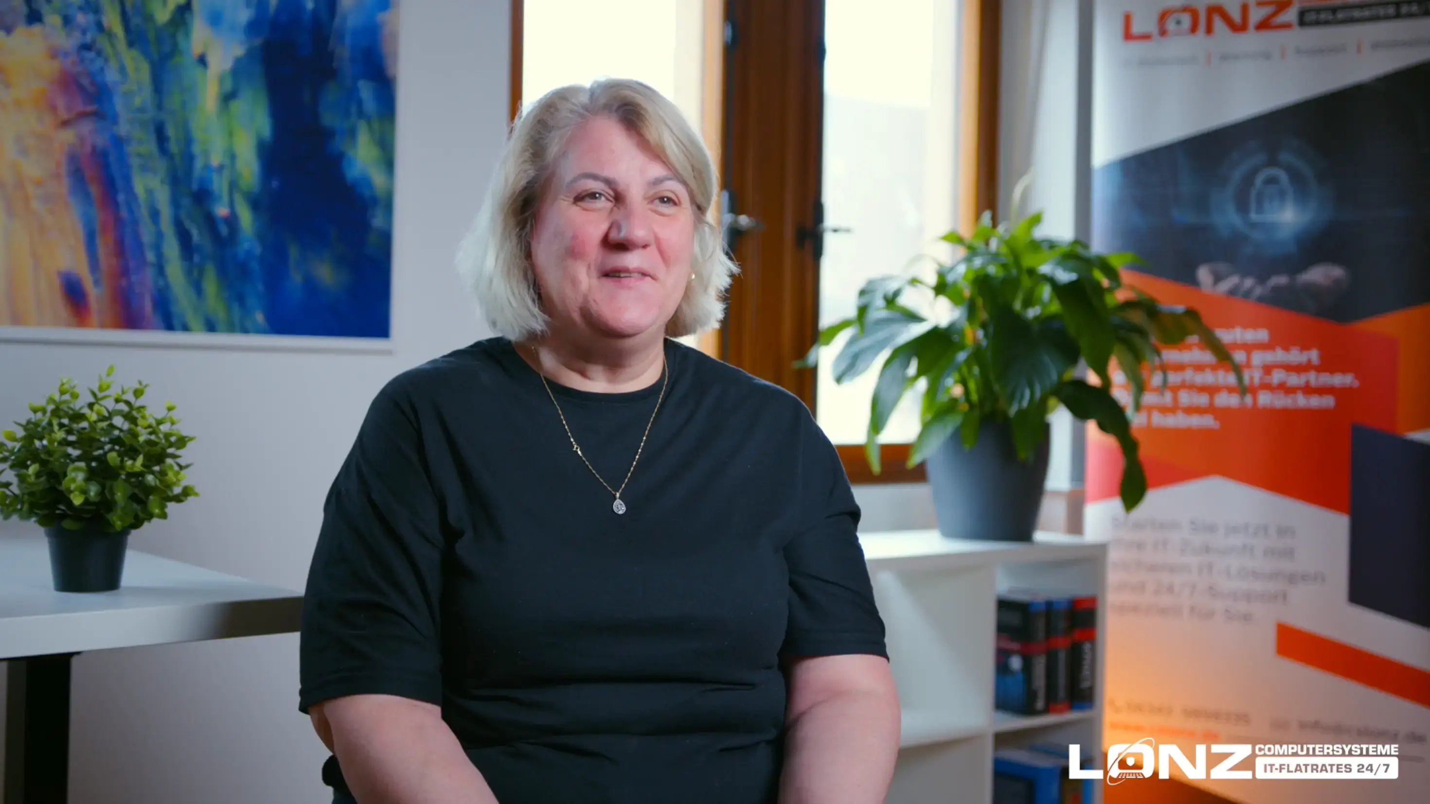 Video-Vorschaubild von Aysel, Mitarbeiterin bei ComputerSysteme Lonz. Sie sitzt in einem Büro und spricht während eines Interviews. Im Hintergrund sind ein Werbebanner und das Logo von Lonz zu sehen.