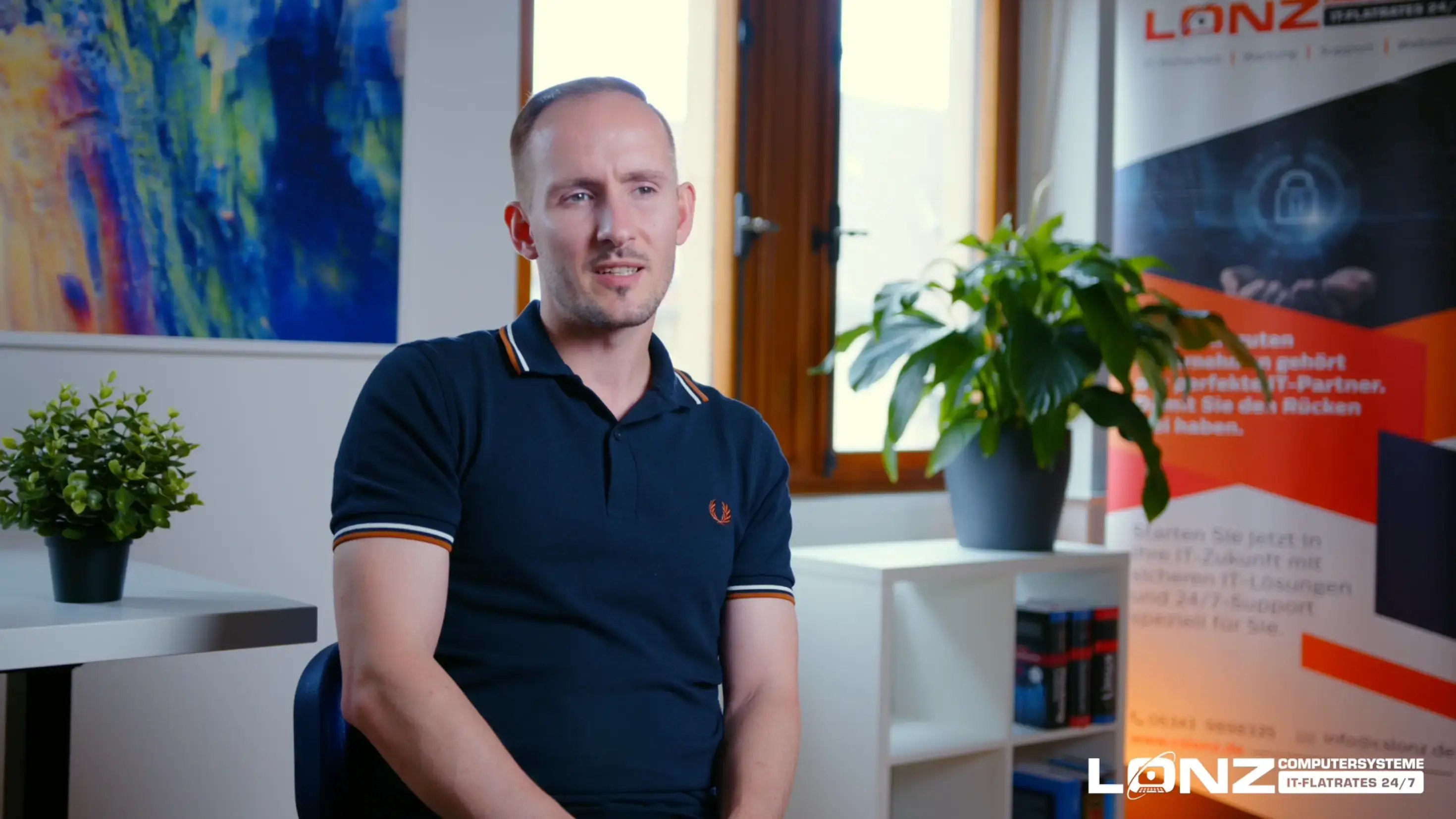 Video-Vorschaubild von Robert, Mitarbeiter bei ComputerSysteme Lonz. Er sitzt in einem Büro und spricht während eines Interviews. Im Hintergrund sind ein Werbebanner und das Logo von Lonz zu sehen.